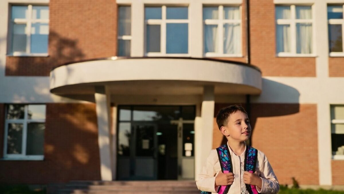 Copil cu ghiozdan în fața unei școli, pregătit pentru înscrierea în clasa pregătitoare